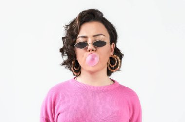 Pembe kazaklı, gözlüklü, beyaz arka planda pembe sakızlı genç bir kadın portresi..