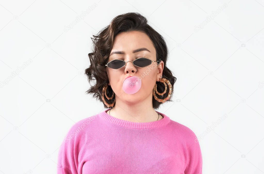 Portrait young woman in a pink sweater with fashion glasses blowing a pink bubble gum on white background.