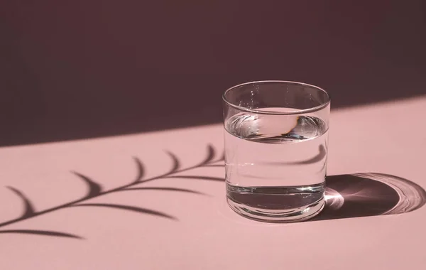 Parlak güneşin aydınlattığı bir bardak su. Pembe zemin üzerinde sıvı bulunan şeffaf bir gemi. Sert gölgeler.