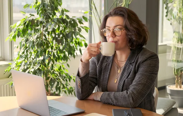 Young business woman having coffee break at work on laptop computer in office. Female freelancer have break at online work and drinking cup of coffee.