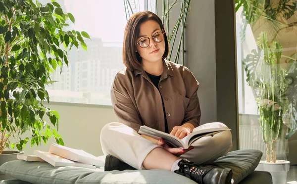 Young woman student reading a book in a bright modern interior. Female with glasses studying. Book club.