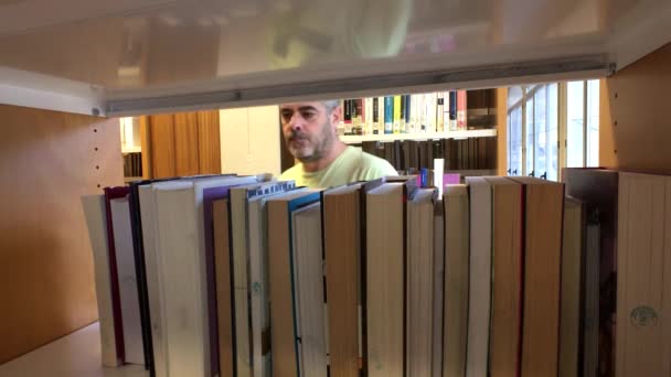 Medelålders man ser ut och plocka upp en bok i ett bibliotek — Stockvideo