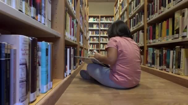 Ung flicka läser en serietidning som sitter på golvet i ett bibliotek — Stockvideo