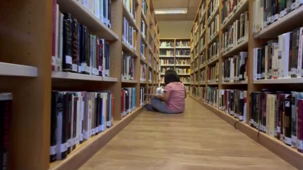 Ung flicka läser en serietidning som sitter på golvet i ett bibliotek — Stockvideo