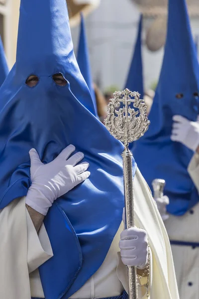 Penitente con túnica de capa blanca y azul mantiene una insignia con escudo de su hermandad durante la estación de penitencia del domingo de Resurrección, toma en Linares, Andalucía, España —  Fotos de Stock