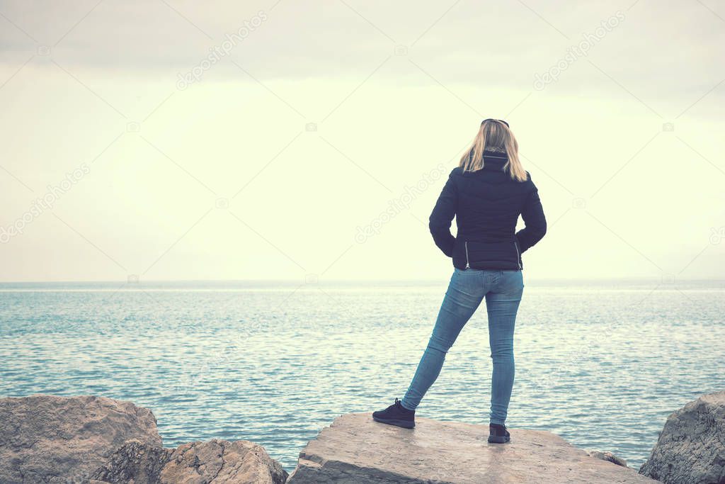 blonde woman looking at the sea in winter season, back view of young lady with blue jeans