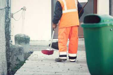 Sokaklarda temizlikçi olarak çalışan bir adam. Kaldırımdaki çöpleri topluyor, yakınlarda bir çöp tenekesi var.)