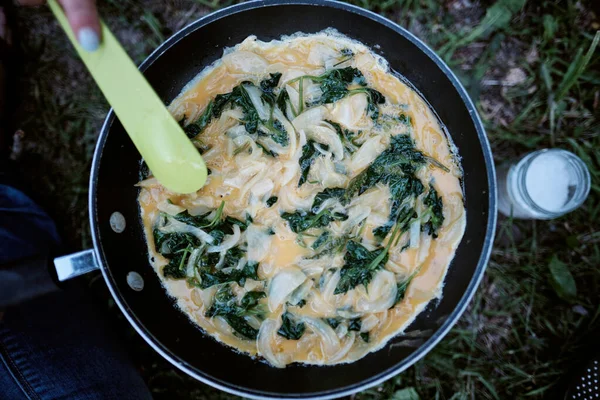 Kamp ateşi kullanarak ıspanaklı ve soğanlı omlet pişiren bir kadın. 