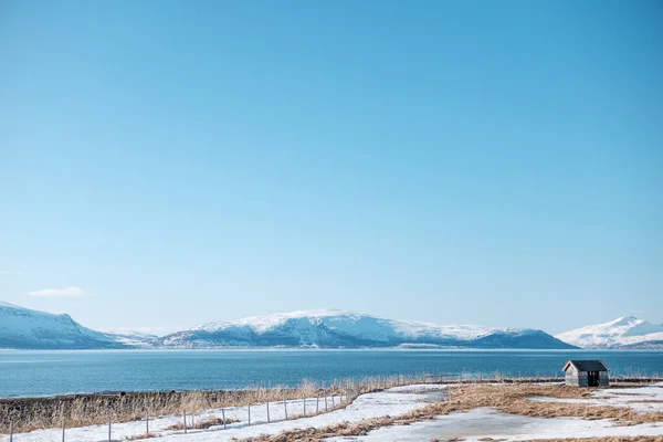 Tromso, Norwegia - 29 kwietnia 2018: Odizolowana opuszczona chatka na brzegu fiordu w słoneczny dzień z górami w tle — Zdjęcie stockowe