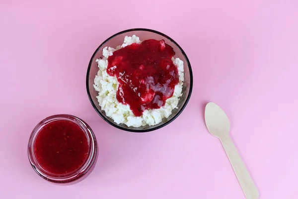 Top view of a bowl with cottage cheese and a glass jar with fresh raspberry jam, accompanied by a wooden spoon. Copy space - the concept of proper nutrition, benefits, natural products — Stock Photo, Image