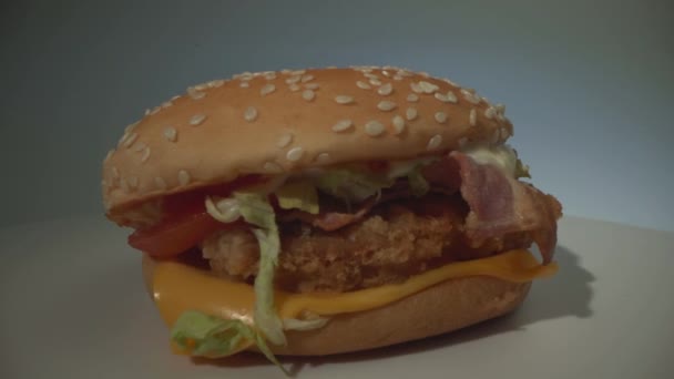 Um delicioso cheeseburger apetitoso com carne, salsicha, tomate, queijo e verduras gira sobre uma superfície branca. Laço — Vídeo de Stock