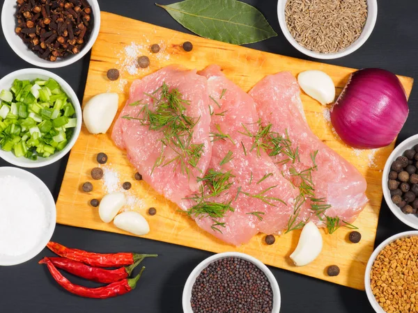 Sliced pieces of meat turkey game filet on a wooden cutting board, garlic, cumin (jeera), salt, fennel, allspice, fenugreek, mustard seeds, oil, green and red onion, cloves on black wooden background. Healthy eating concept