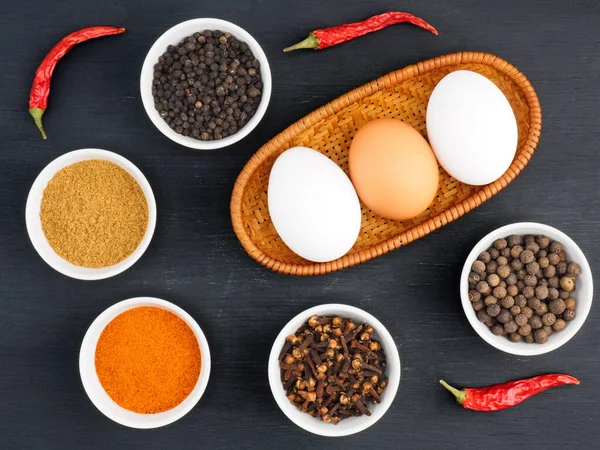 Raw chicken eggs, black pepper, masala powder, cloves, allspice, red chili pepper, black pepper on black wooden background. Ingredients for making scrambled eggs or omelet. Organic food concept