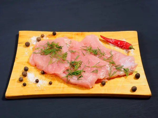 Sliced pieces of meat turkey game filet, fennel, allspice, red chili pepper, salt on a wooden cutting board on black wooden background. Healthy eating concept