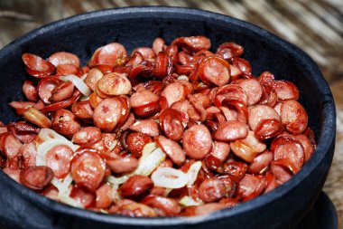 Kızarmış et sosisi, soğan ve baharat, ahşap masada, siyah kil tabakta.