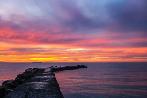 A pier on the Mediterranean Sea in Tuscany at sunset with an island and a sailboat in the background