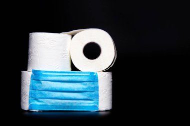 Siyah arka planda mavi tıbbi maskeli beyaz tuvalet kağıdı ruloları var. Coronavirüs salgını panik konsepti