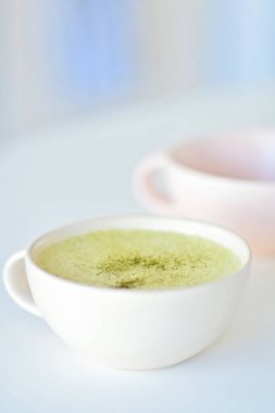 Beyaz bir masada kibrit sütlü beyaz fincan, arka planda açık pembe bardak, fincanda yeşil çay latte. Üst Manzara. Uzayı kopyala Yeşil çay, matcha latte enerji takviyesi ve matcha 'nın sağlıklı faydalarından zevk almanın lezzetli bir yoludur. Matcha bir p
