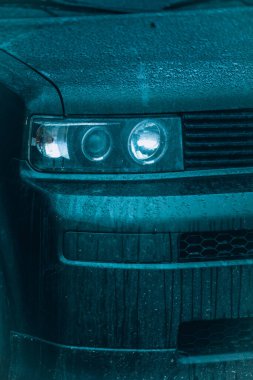 Moda arabası. Yağmur damlaları içinde modern koyu renkli bir arabanın önü. Farlar ve kaput yakın çekim.