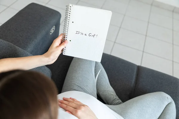 Mladá těhotná žena drží zápisník sedící na pohovce - G — Stock fotografie