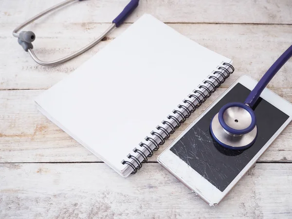 Smartphone rotto con stetoscopio e notebook su dorso in legno — Foto Stock