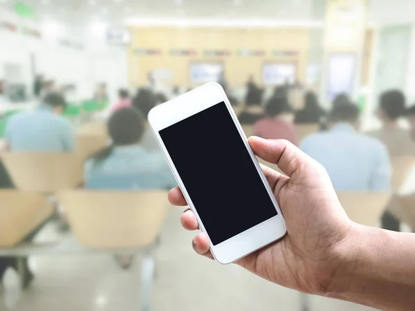 Hand holding mobile phone over blurred people sitting at bank — Stock Photo, Image