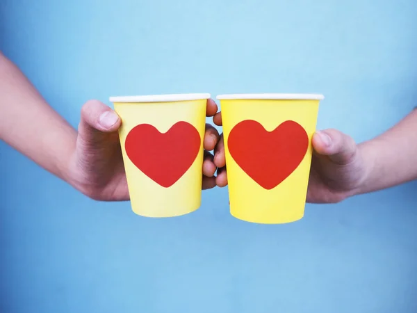 Manos sosteniendo copas amarillas con forma de corazón rojo sobre pared azul . —  Fotos de Stock
