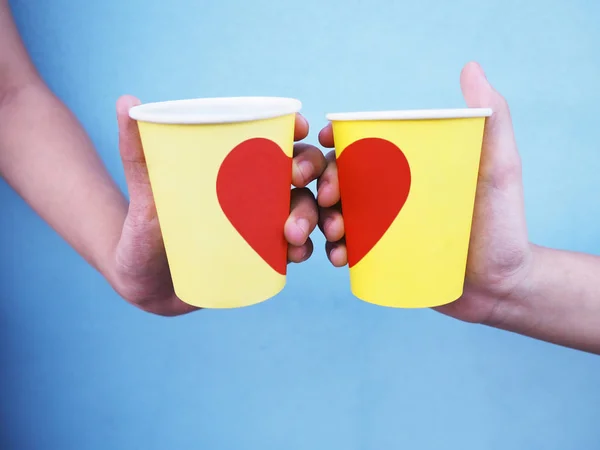 Manos sosteniendo copas amarillas con forma de corazón rojo sobre pared azul . —  Fotos de Stock