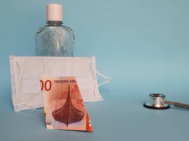 Norveç banknotu 100 kron, yüz maskesi, jel alkollü şişe ve mavi arka planda stetoskop.