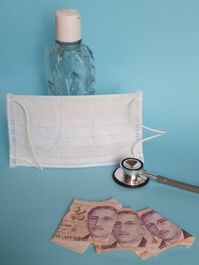 İki dolarlık Singapur banknotları, yüz maskesi, jel alkollü şişe ve mavi arka planda steteskop.