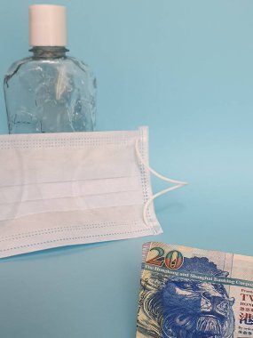 Hong Kong banknotu 20 dolar, yüz maskesi, jel alkollü şişe ve mavi arka plan.