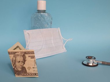 20 dolarlık Amerikan banknotu, maske, jel alkollü şişe ve mavi arka planda steteskop.