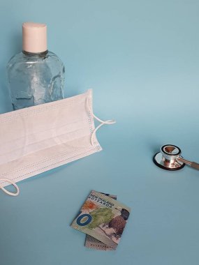 Yeni Zelanda banknotu 10 dolar, maske, jel alkollü şişe ve mavi arka planda stetoskop.