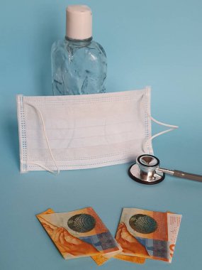 İsviçre banknotları 10 frank, maske, jel alkollü şişe ve mavi arka planda steteskop.