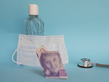 20 kronluk banknot, maske, jel alkollü şişe ve mavi arka planda stetoskop.