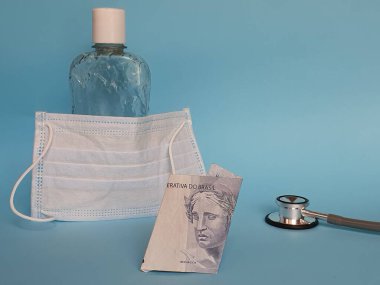 İki reais Brezilya banknotu, maske, jel alkollü şişe ve mavi arka planda steteskop.