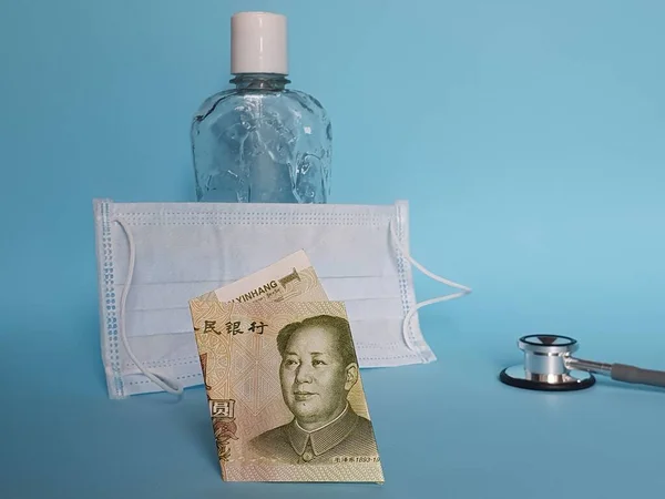 chinese banknote of one yuan, mask, bottle with gel alcohol and stethoscope on blue background