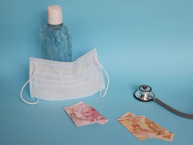 Türk banknotları, yüz maskesi, jel alkollü şişe ve mavi arka planda steteskop.