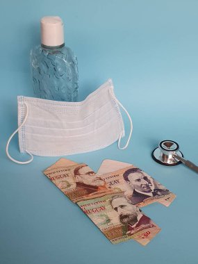 uruguay banknotları, yüz maskesi, jel alkollü şişe ve mavi arka planda steteskop.