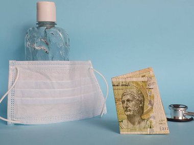 Tunus banknotu beş dinar, yüz maskesi, jel alkollü şişe ve mavi arka planda stetoskop.