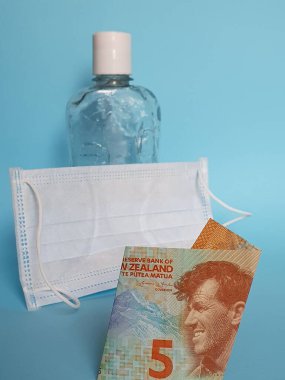 Yeni Zelanda 'da beş dolarlık banknot, yüz maskesi, jel alkollü şişe ve mavi arka plan.