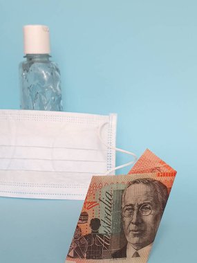 20 dolarlık Avustralya banknotu, yüz maskesi, jel alkollü şişe ve mavi arka plan.