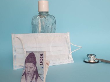 Güney Kore banknotu 1000 won, maske, jel alkollü şişe ve mavi arka planda steteskop.