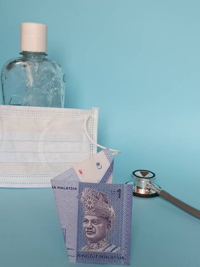 Bir halka, maske, jel alkollü şişe ve mavi arka planda stetoskop içeren Malezya banknotu