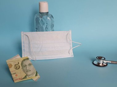 Beş dolarlık Singapur banknotu, maske, jel alkollü şişe ve mavi arka planda steteskop.