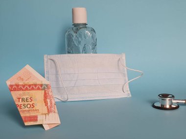 Üç pezoluk Küba banknotu, maske, jel alkollü şişe ve mavi arka planda steteskop.