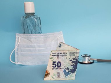Arjantin banknotu 50 peso, maske, jel alkollü şişe ve mavi arka planda stetoskop.