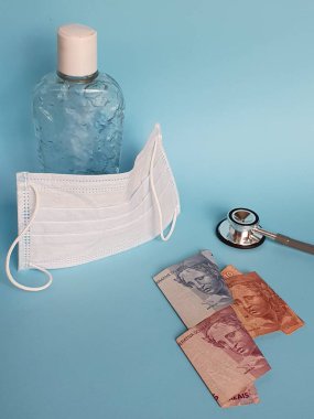 Brezilya banknotları, maske, jel alkollü şişe ve mavi arka planda steteskop.