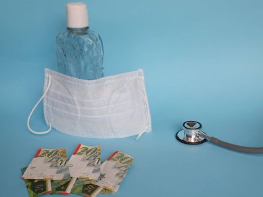 İsrail banknotları 20 şekel, maske, jel alkollü şişe ve mavi arka planda steteskop