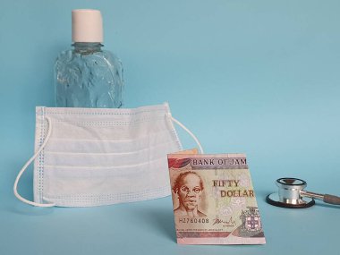 50 dolarlık Jamaika banknotu, maske, jel alkollü şişe ve mavi arka planda steteskop.